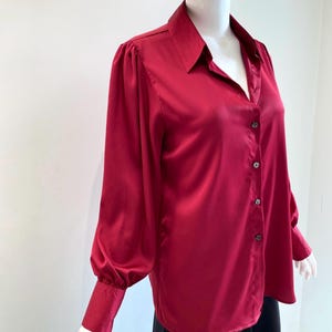 May include: A deep red satin blouse with a classic collar and button-down front. The long sleeves feature a gathered detail at the shoulder and a buttoned cuff. The blouse is displayed on a mannequin.