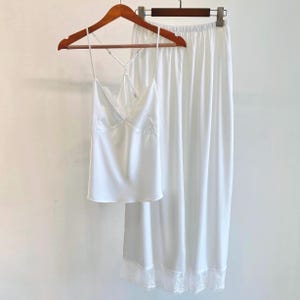 May include: A white satin pyjama set, comprising a camisole with lace trim and spaghetti straps, and a matching skirt with a lace hem. The set is displayed on a wooden hanger against a plain white backdrop.