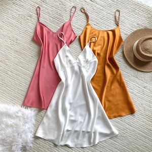 May include: Three sleeveless slip dresses in solid satin fabric, featuring thin spaghetti straps and V-necklines. The dresses are presented in pink, white, and mustard yellow colors, arranged alongside a straw hat with a white ribbon band.