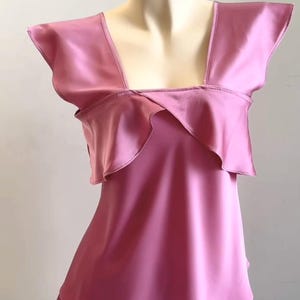 May include: A pink satin pajama set displayed on a mannequin. The top features a square neckline and ruffled detailing. The shorts have a layered design, creating a flowing silhouette. The fabric has a shiny, smooth texture.