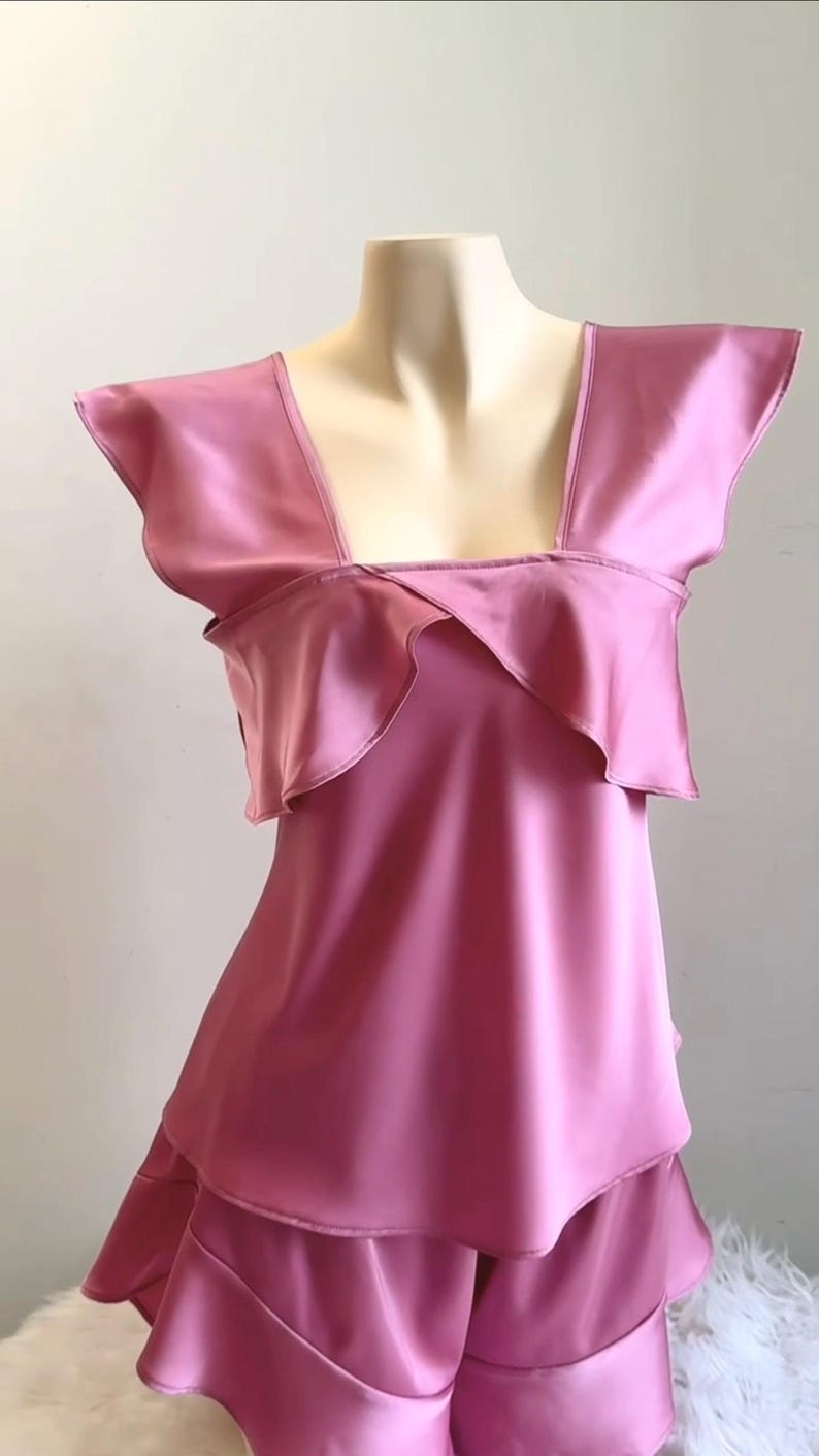 May include: A pink satin pajama set displayed on a mannequin. The top features a square neckline and ruffled detailing. The shorts have a layered design, creating a flowing silhouette. The fabric has a shiny, smooth texture.