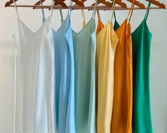 Könnte beinhalten: Eine Kollektion von sechs ärmellosen Trägerkleidern an Holzkleiderbügeln, angeordnet in einem Farbverlauf von Weiß über Hellblau, Minzgrün, Blassgelb und Goldorange bis hin zu Smaragdgrün, jeweils mit dünnen Spaghettiträgern versehen.