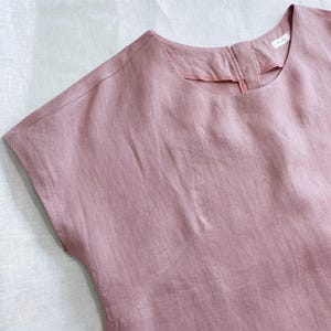 May include: A dusty rose-colored top with short sleeves, a round neckline, and a visible seam running diagonally across the chest. The garment features a small white label inside the back collar area and has a textured, woven fabric appearance.
