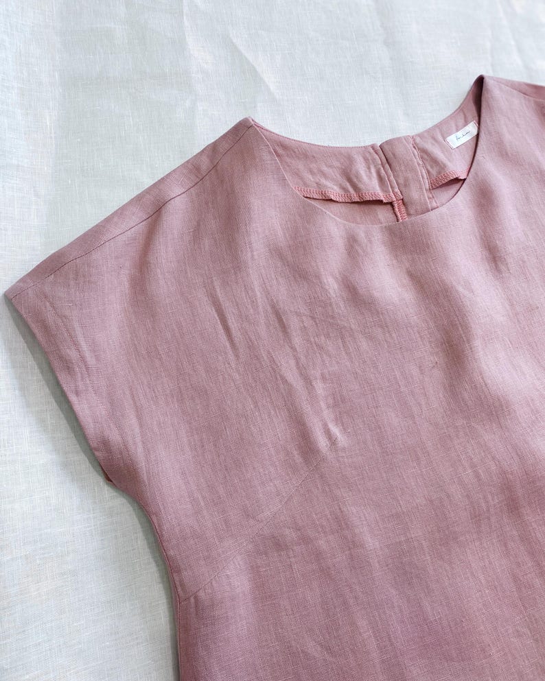 May include: A dusty rose-colored top with short sleeves, a round neckline, and a visible seam running diagonally across the chest. The garment features a small white label inside the back collar area and has a textured, woven fabric appearance.