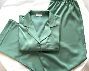Könnte beinhalten: Ein salbeigrüner Pyjama-Set bestehend aus einem langärmeligen Hemd mit Knopfleiste und Reverskragen, einer Brusttasche sowie einer passenden weit geschnittenen Hose mit elastischem Bund. Der Stoff hat eine glatte, satinartige Oberfläche.