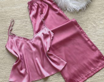 Könnte beinhalten: Ein zweiteiliges Schlafanzug-Set aus rosa Satin. Das Set besteht aus einem Top mit dünnen Trägern und einem gerüschten Ausschnitt sowie einer Hose mit weitem Bein. Der Stoff hat eine glänzende, glatte Textur.
