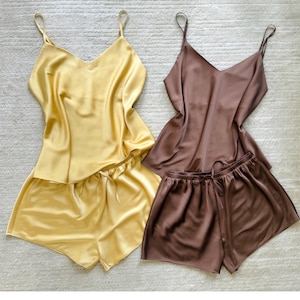 Könnte beinhalten: Zwei Sets aus Satin-Nachtwäsche. Ein Set ist goldgelb, das andere ist ein gedämpftes Braun. Jedes Set enthält ein Camisole-Top mit dünnen Trägern und V-Ausschnitt sowie passende Shorts mit Kordelzug. Der Stoff hat eine glänzende, glatte Textur.