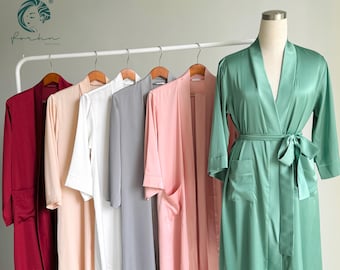 Könnte beinhalten: Sieben Satin-Bademäntel hängen an einem Gestell. Die Bademäntel sind in verschiedenen Farben, darunter Burgunderrot, Blush, Weiß, Grau und Rosa. Die Bademäntel haben lange Ärmel und einen Bindegürtel in der Taille.