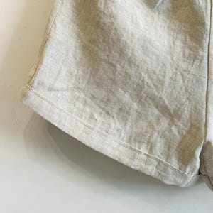 May include: Close-up of light beige linen shorts. The shorts have a relaxed fit with a drawstring waistband and pockets. The fabric has a textured appearance, and the stitching is visible along the edges.