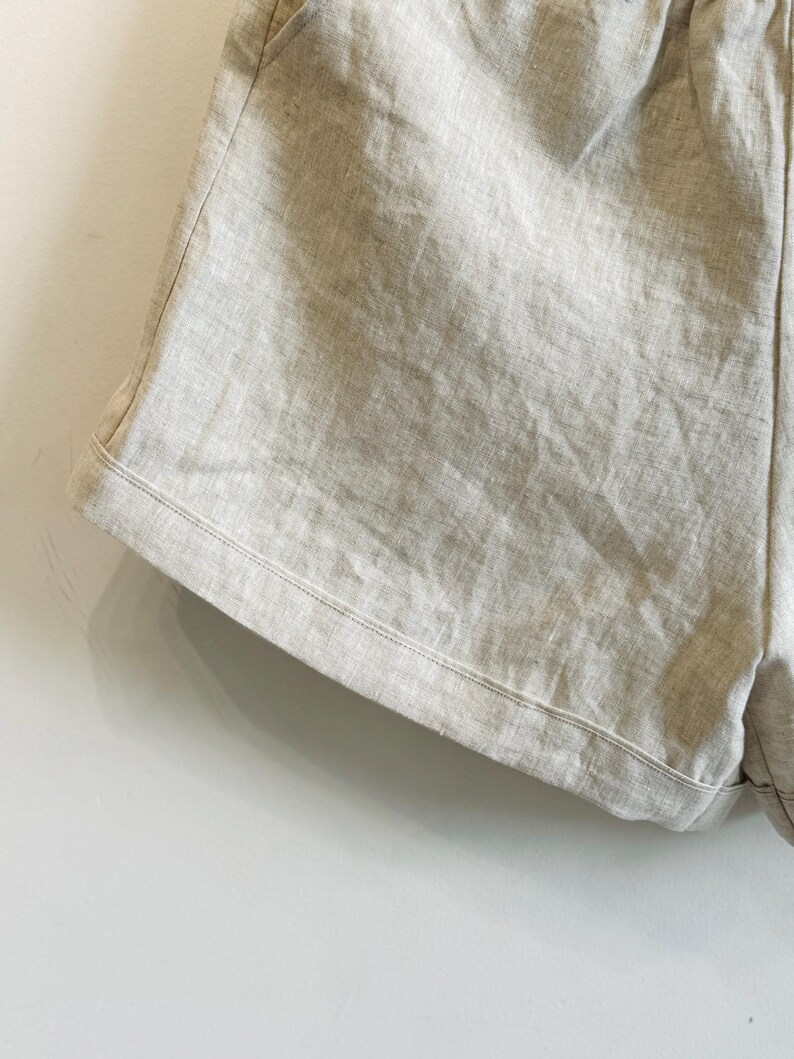 May include: Close-up of light beige linen shorts. The shorts have a relaxed fit with a drawstring waistband and pockets. The fabric has a textured appearance, and the stitching is visible along the edges.