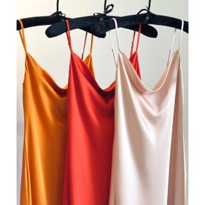 Może przedstawiać: Trzy jedwabne sukienki w kolorach pomarańczowym, czerwonym i różowym wiszą na czarnych aksamitnych wieszakach. Sukienki mają dekolt w kształcie łódki i cienkie ramiączka spaghetti, prezentując minimalistyczny design. Gładka, zwiewna tkanina sugeruje luksusowy charakter.
