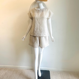 May include: A beige linen shorts set and a straw hat with a white band on a mannequin. The shorts are high-waisted and the top has short sleeves. The hat has a wide brim, perfect for summer.