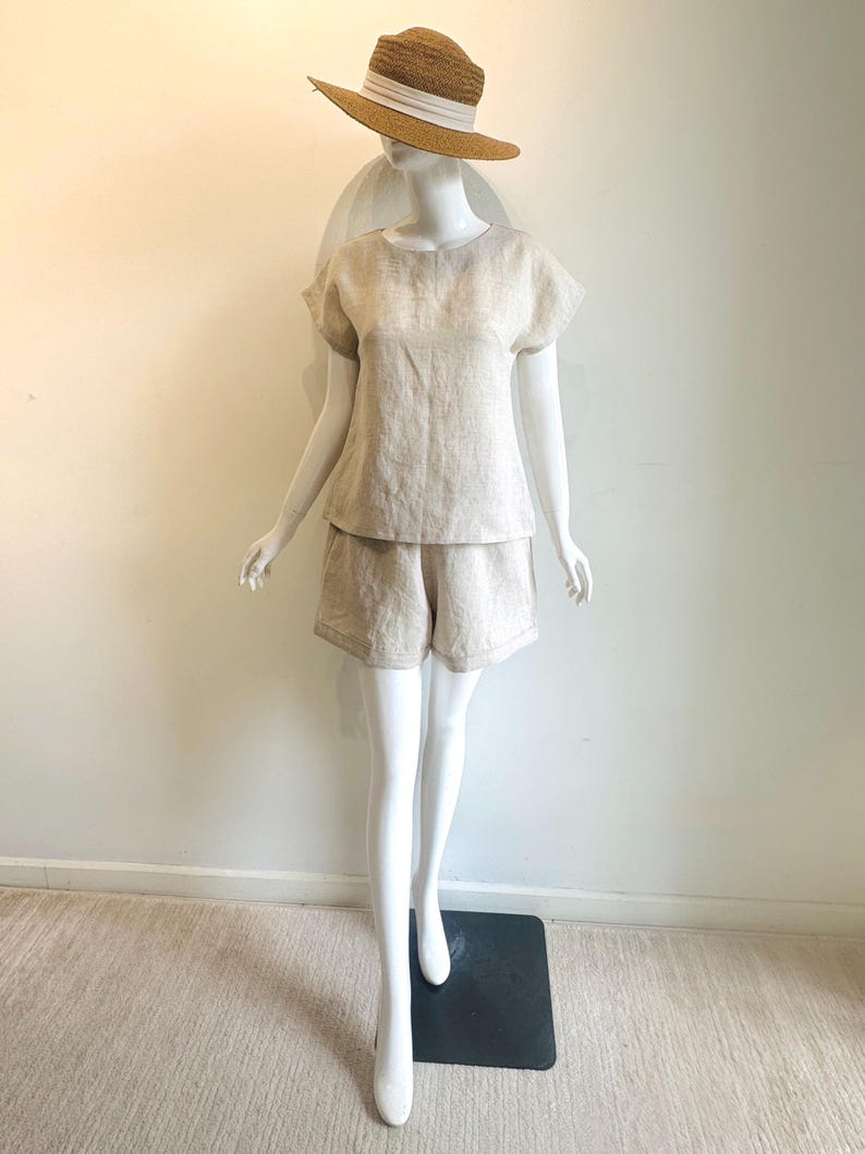May include: A beige linen shorts set and a straw hat with a white band on a mannequin. The shorts are high-waisted and the top has short sleeves. The hat has a wide brim, perfect for summer.