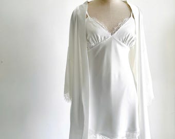 Könnte beinhalten: Ein Set bestehend aus einem weißen Satin-Nachthemd und einem passenden Morgenmantel. Das Nachthemd hat einen V-Ausschnitt mit zarter Spitzenborte. Der Morgenmantel hat lange Ärmel mit Spitzenbesatz. Beide Teile sind aus glattem, weißem Stoff.