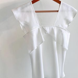 May include: A white sleeveless top with a square neckline and ruffled detailing. The top is hanging on a wooden hanger against a white background. The fabric appears to be lightweight and flowy.
