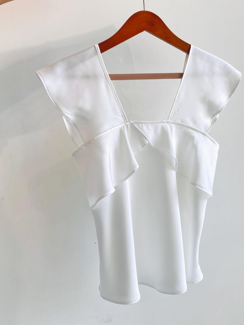 May include: A white sleeveless top with a square neckline and ruffled detailing. The top is hanging on a wooden hanger against a white background. The fabric appears to be lightweight and flowy.