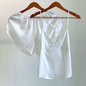 May include: A white satin pajama set, including shorts and a camisole top, hangs on a wooden hanger. The camisole features a V-neck design and spaghetti straps. The shorts have a relaxed fit. The set is made of a smooth, shiny fabric.