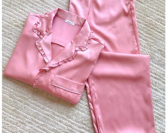 Puede incluir: Un conjunto de pijama rosa claro que incluye una camisa de manga larga con botones, cuello con volantes y un bolsillo en el pecho, junto con pantalones largos a juego. La camisa tiene una pequeña etiqueta blanca en el cuello con texto negro.