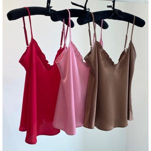 May include: Three silk camisole tops in red, pink, and brown hang on a black velvet hanger. Each top has thin spaghetti straps and a ruffled neckline. The tops are made of a smooth, flowing fabric.