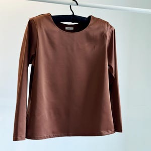 May include: A long-sleeved, brown shirt with a round neckline hangs on a black hanger. The shirt is made of a smooth fabric and appears to be a solid color. The shirt is displayed against a white background.