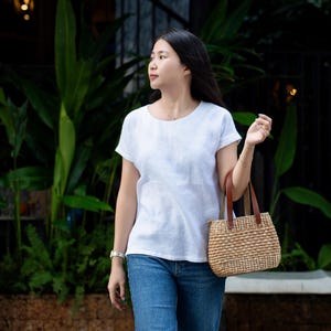 May include: A white short-sleeved crew neck top paired with blue denim jeans and a woven straw handbag featuring brown leather handles. The handbag has a structured rectangular base and a textured, natural fiber exterior with a zipper closure.