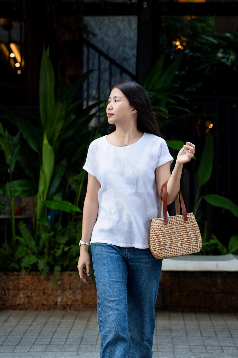 May include: A white short-sleeved crew neck top paired with blue denim jeans and a woven straw handbag featuring brown leather handles. The handbag has a structured rectangular base and a textured, natural fiber exterior with a zipper closure.