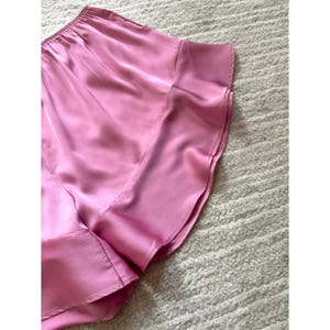 May include: A close-up of a pink satin garment, possibly a top or blouse, with a flowing, draped design. The fabric has a subtle sheen, and the garment appears to be lightweight. The image is taken from an angle, showing the texture of the fabric.