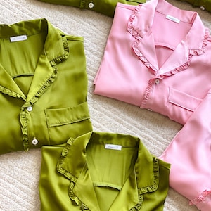 May include: Overhead shot of folded satin pyjamas in shades of green and pink. The pyjama tops have a ruffled collar and pocket. The pyjamas are neatly arranged on a soft, textured surface. Each top has a small tag.