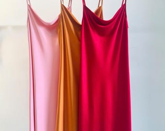 Könnte beinhalten: Drei ärmellose Slip-Kleider hängen an Holzkleiderbügeln. Die Kleider haben einen Wasserfall-Ausschnitt und dünne Spaghettiträger. Die Farben von links nach rechts sind Hellrosa, Goldgelb und leuchtendes Magenta. Jedes Kleid hat eine glatte Oberfläche.