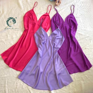 May include: Four satin slip dresses in shades of pink, purple, and lavender are arranged on a white surface. The dresses have a V-neckline and spaghetti straps. The logo for "Forhn" is in the upper left corner.
