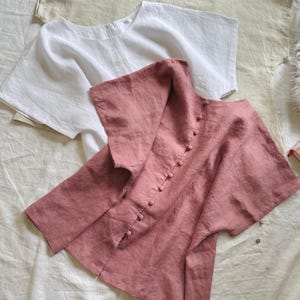 May include: Three short-sleeved blouses in beige, white, and dusty rose. The dusty rose blouse features a vertical row of matching fabric-covered buttons down the center. Each blouse has a wide, loose fit with a rounded neckline and short sleeves.
