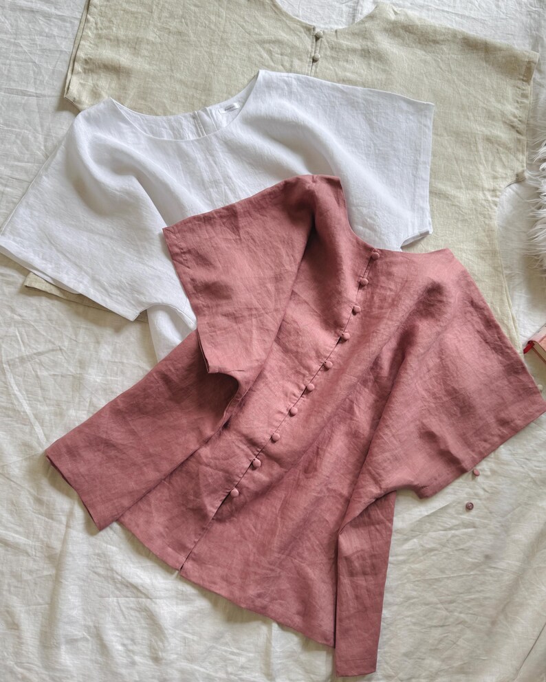 May include: Three short-sleeved blouses in beige, white, and dusty rose. The dusty rose blouse features a vertical row of matching fabric-covered buttons down the center. Each blouse has a wide, loose fit with a rounded neckline and short sleeves.