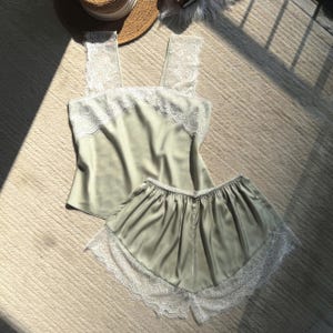 Puede incluir: Conjunto de pijama de satén verde salvia con ribete de encaje blanco. El conjunto incluye una camiseta sin mangas con tirantes anchos y pantalones cortos. Un sombrero de paja es visible en el fondo. El conjunto está hecho de un material suave y sedoso.