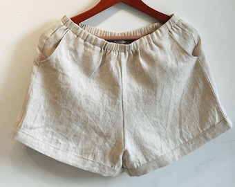 May include: Beige shorts featuring an elasticated waistband, two side pockets, and a cuffed hem. The garment is constructed from a textured fabric with a visible weave, hanging from a wooden hanger against a plain white background.