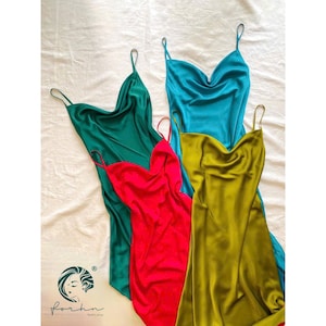 Könnte beinhalten: Vier Satin-Slipkleider in Türkis, Grün, Rot und Olivgrün sind auf einer weißen Oberfläche angeordnet. Jedes Kleid hat einen Wasserfallausschnitt und Spaghettiträger. Die Kleider sind von der Marke "forhn".