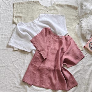 May include: Three short-sleeved shirts in beige, white, and dusty rose. Each shirt features a round neckline, a loose fit, and a visible button closure at the back of the collar. The fabric has a textured appearance, suggesting a natural weave.