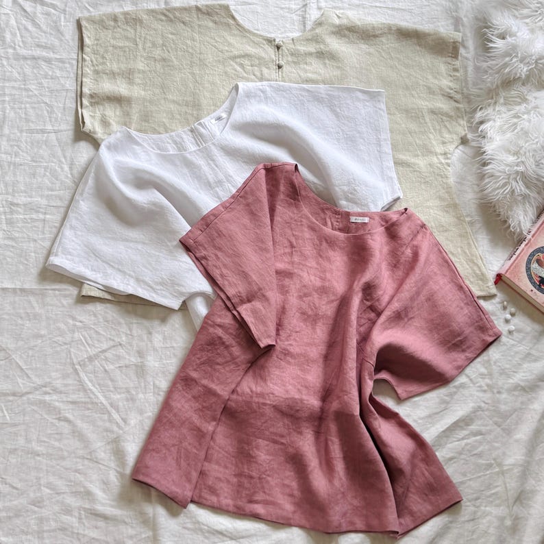 May include: Three short-sleeved shirts in beige, white, and dusty rose. Each shirt features a round neckline, a loose fit, and a visible button closure at the back of the collar. The fabric has a textured appearance, suggesting a natural weave.