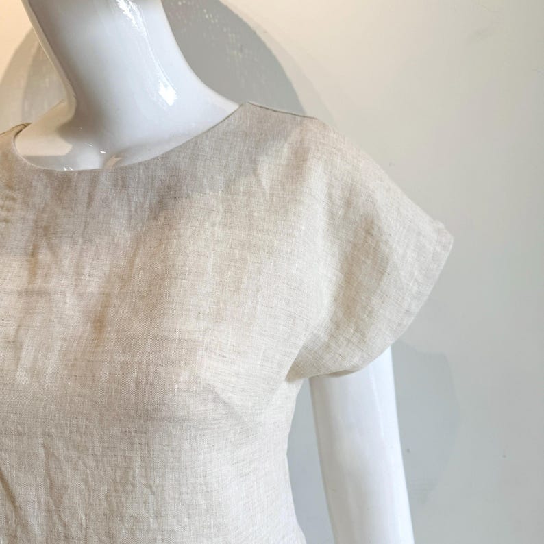 May include: A beige linen top with short sleeves and a round neckline. The fabric has a textured appearance. The top is displayed on a white mannequin.