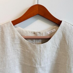 May include: A light beige linen top with a scoop neckline, hanging on a wooden hanger. The top has a small tag with text and the size XS. The fabric appears to be textured and lightweight.