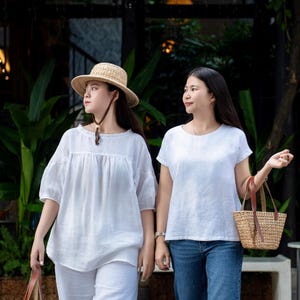 May include: Two white linen tops, one with a gathered neckline and puff sleeves, the other with a round neck and short sleeves. One top is paired with white linen trousers and a straw hat, the other with blue denim jeans. Both carry woven straw bags.