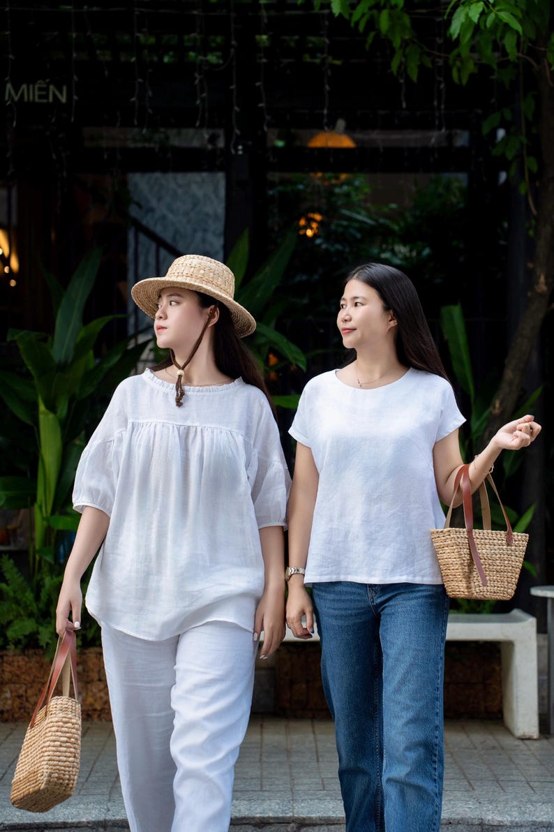 May include: Two white linen tops, one with a gathered neckline and puff sleeves, the other with a round neck and short sleeves. One top is paired with white linen trousers and a straw hat, the other with blue denim jeans. Both carry woven straw bags.