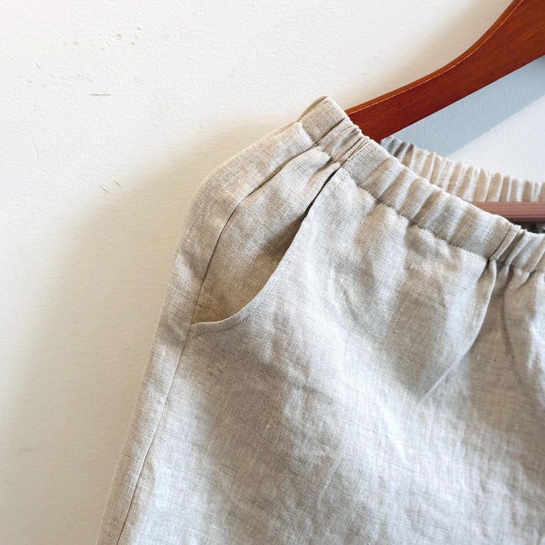 May include: Close-up of light beige linen trousers. The trousers feature an elasticated waistband and a side pocket. The fabric appears lightweight and breathable, suitable for warm weather. The trousers are hanging on a wooden hanger.