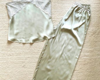 Könnte beinhalten: Ein Pyjama-Set aus salbeigrünem Satin, bestehend aus einem Camisole mit weißer Spitzenborte an den Trägern und am Ausschnitt sowie einer passenden weiten Hose mit weißer Spitzenborte am Saum, ergänzt durch einen Strohhut mit weißem Band.