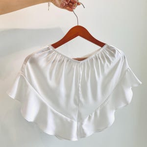 May include: A white, ruffled top with a scalloped neckline, hanging on a wooden hanger. The top has a flowing, silky appearance and a V-neck design. The ruffles add a decorative element to the garment.