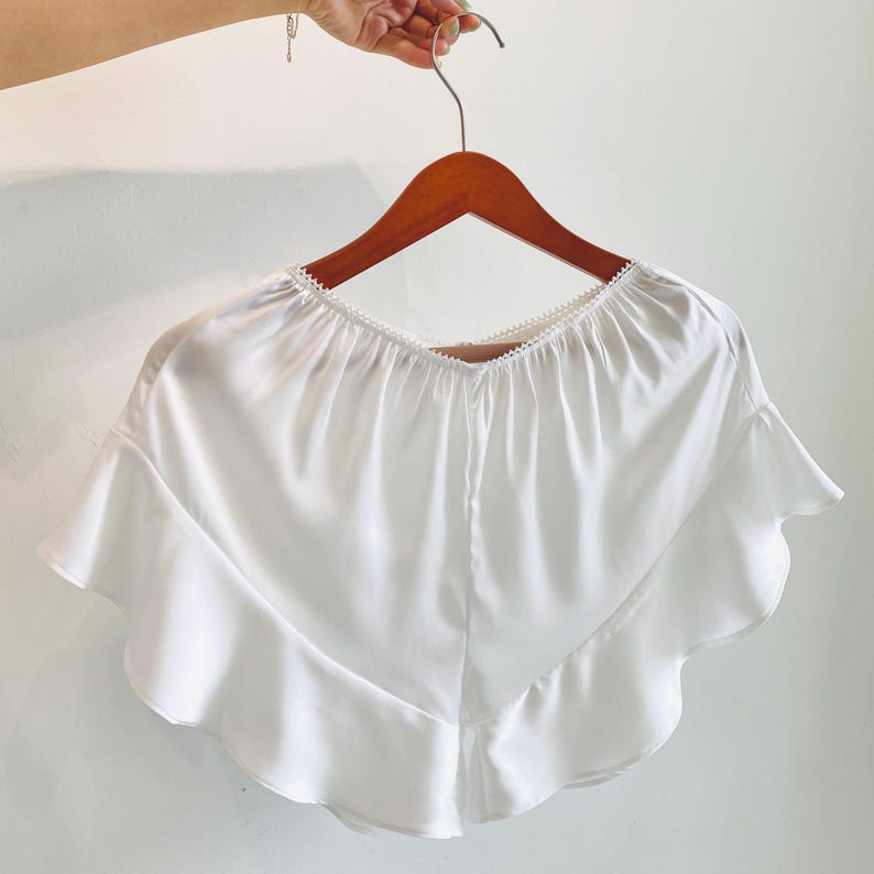 May include: A white, ruffled top with a scalloped neckline, hanging on a wooden hanger. The top has a flowing, silky appearance and a V-neck design. The ruffles add a decorative element to the garment.