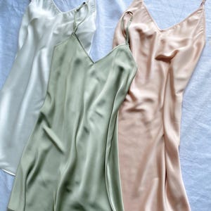 K&ouml;nnte beinhalten: Drei seidene Schlupfkleider in Wei&szlig;, Salbeigr&uuml;n und Blush-Rosa. Die Kleider haben d&uuml;nne Tr&auml;ger und einen V-Ausschnitt.