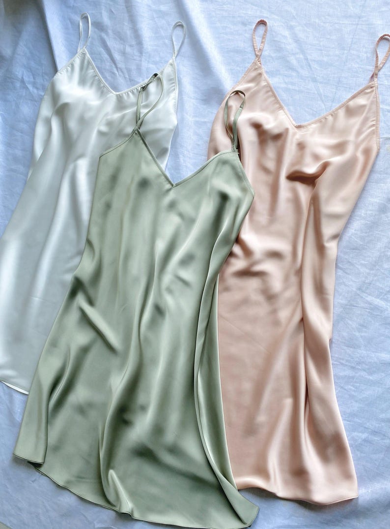 K&ouml;nnte beinhalten: Drei seidene Schlupfkleider in Wei&szlig;, Salbeigr&uuml;n und Blush-Rosa. Die Kleider haben d&uuml;nne Tr&auml;ger und einen V-Ausschnitt.
