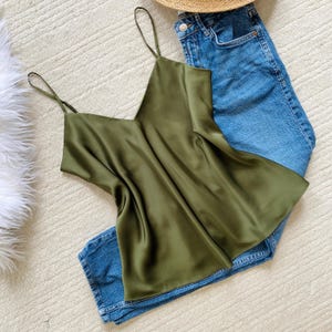 May include: An olive green satin camisole with thin spaghetti straps and a V-neckline, paired with blue denim jeans featuring a silver button and a straw hat brim visible at the top edge of the frame.