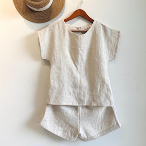 May include: A beige linen two-piece set, comprising a short-sleeved top and matching shorts, hangs on a wooden hanger. A straw hat with a white band is partially visible in the background. The set is made of a lightweight, textured fabric.