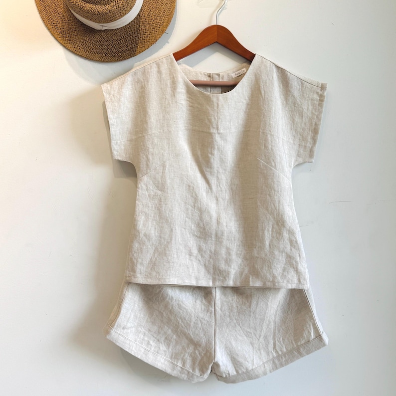 May include: A beige linen two-piece set, comprising a short-sleeved top and matching shorts, hangs on a wooden hanger. A straw hat with a white band is partially visible in the background. The set is made of a lightweight, textured fabric.